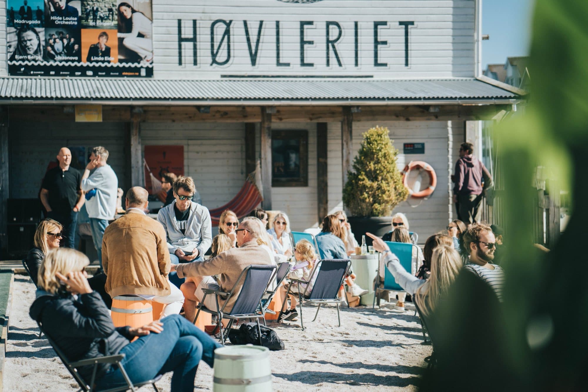 Solrik strandfest på sanden foran det hvite Høvleriet-bygget, med gjester i fluktstoler, hengekøye og tønnebord.