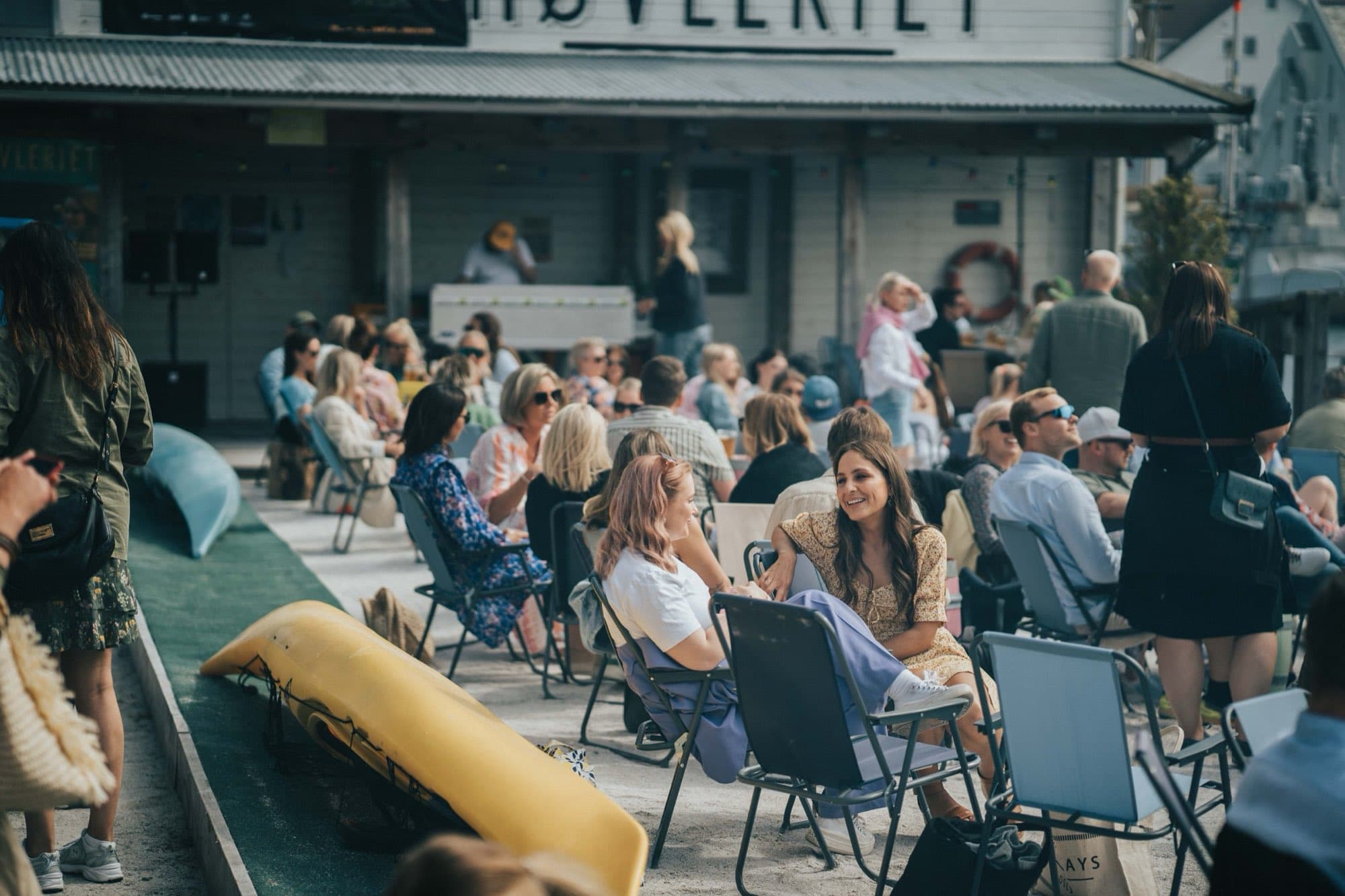 Fullsatt sandstrand med solstoler og gul kajakk foran det hvite Høvleriet-bygget en solrik dag.