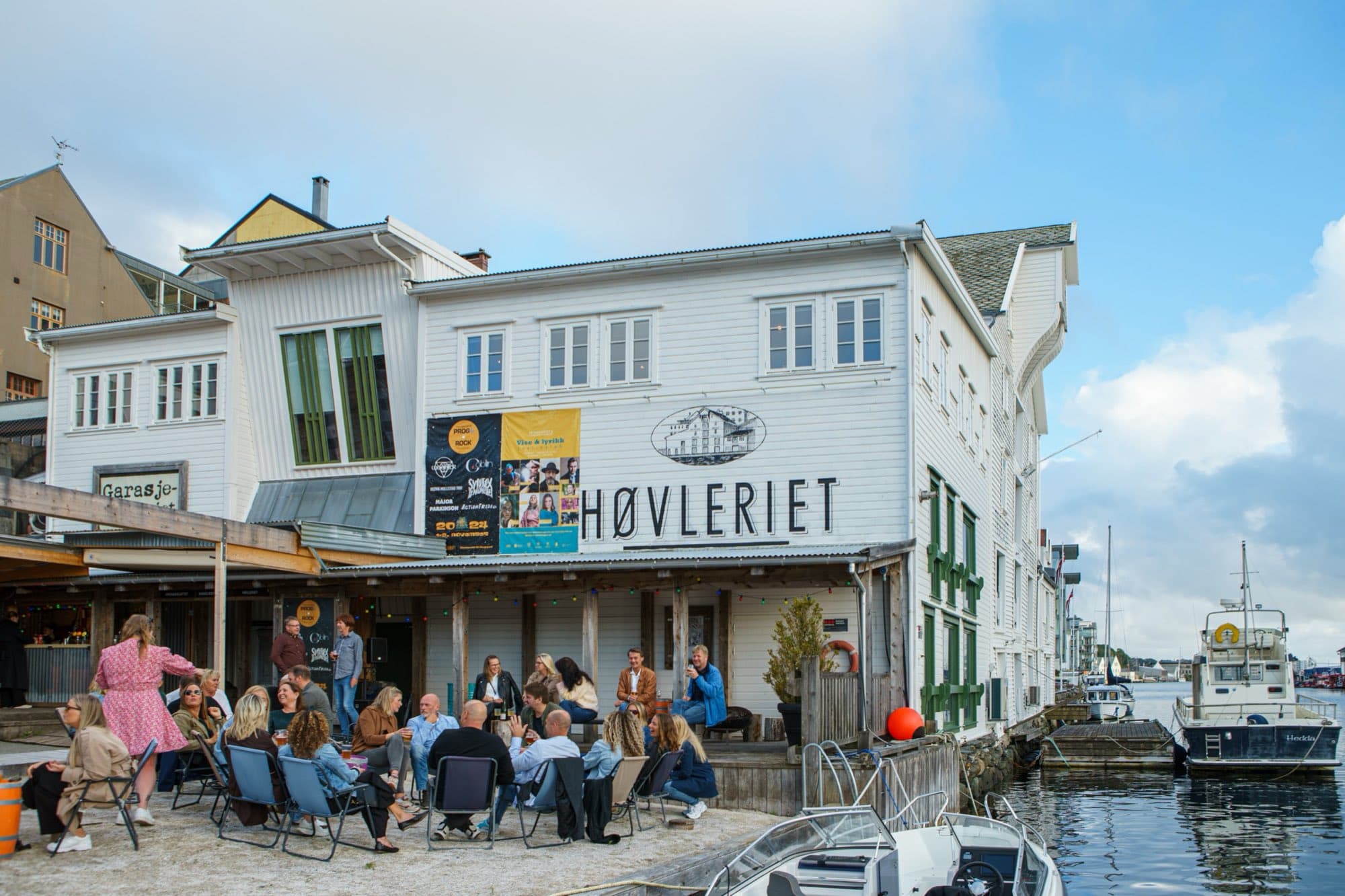 Høvleriet i sol med festivalplakater på fasaden og gjester i campingstoler på sanden ved kaikanten.