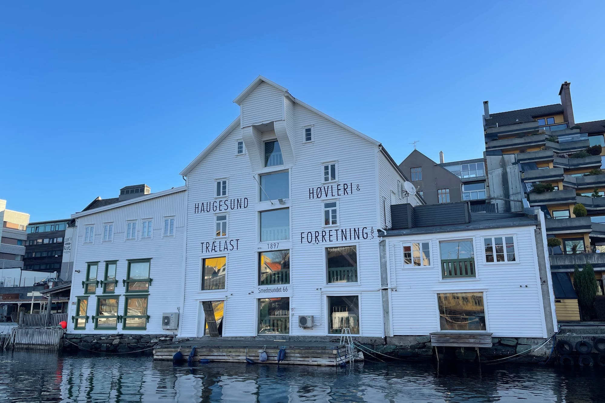 Høvleriet sett fra sjøen — den hvite 1897-fasaden i tre etasjer med «Haugesund Trælast» og «Høvleri & Forretning» malt over veggen, og flytebrygge langs vannlinjen.