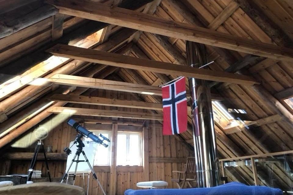 Loftet i Vindkjeften under åpne takstoler, med teleskop ved vinduet, norsk flagg på skorsteinen og gyngestol.