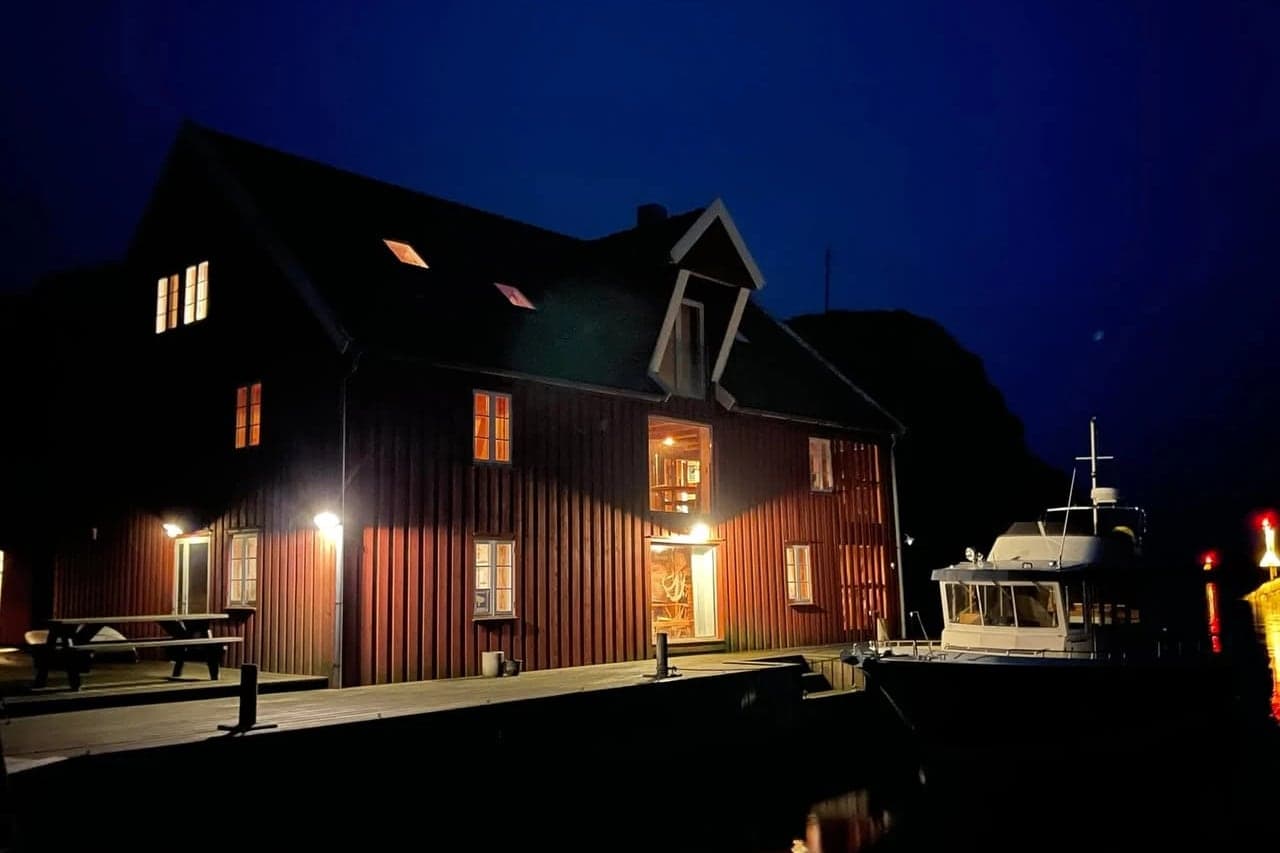 Vindkjeften opplyst i blåtimen, med lys i vinduene, lykter langs kaia og en båt fortøyd i mørket.