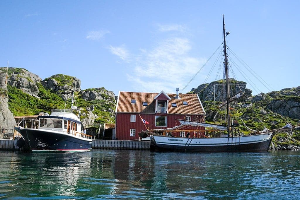 Vindkjeften i sommersol med en seilskute og en motorbåt til kai, og bratte knauser bak huset.