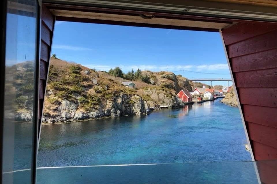 Utsyn gjennom den åpne kaidøra i Vindkjeften mot sundet, med grønne knauser, røde sjøhus og brua i det fjerne.