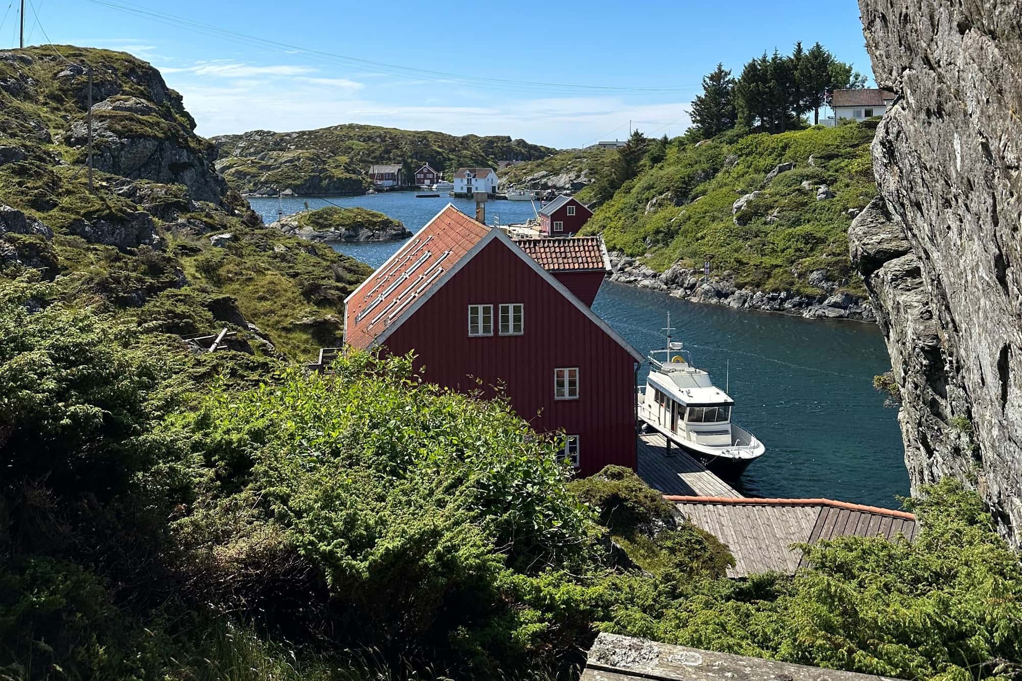 Det røde sjøhuset Vindkjeften ved sundet på Røvær, med båt ved kaien, holmer rundt og bebyggelsen på øya bak.